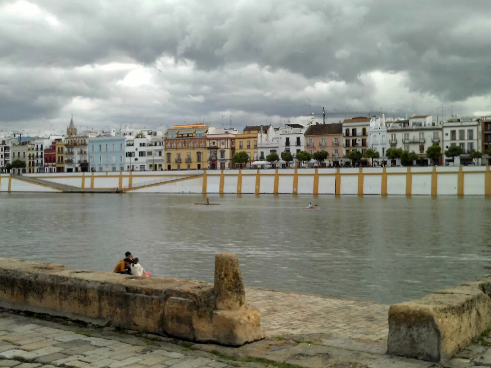 The white and coloured houses of Calle Betis along the river, seen from the opposite bank under a cloudy sky